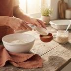A person using a digital measuring spoon to scoop ingredients over a wooden kitchen surface with baking supplies in the backg