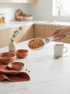 A person holding a digital measuring spoon with an LCD display over a marble countertop with terracotta kitchen accessories.